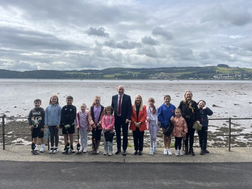 A group of children and two adults standing along a waterfront promenade with hills and water in the background, posing for a photo during a community outing.