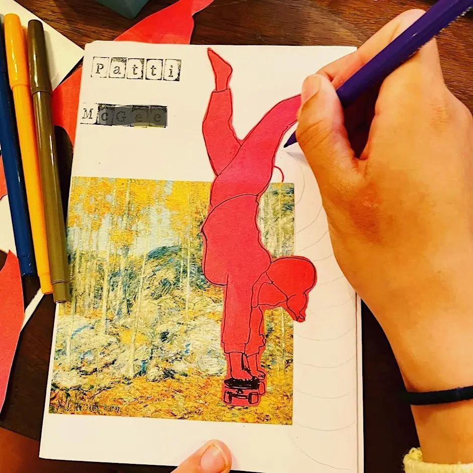 A close-up, top-down view of a person’s hand using a purple pencil to draw on a handmade zine. The zine features a collage of a person in a red outfit performing a handstand over an autumnal landscape. Coloured pens and paper scraps are scattered on the wooden table nearby.