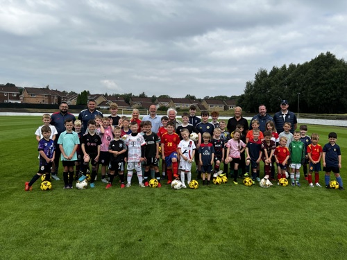 Children and adults posing together on a grassy football pitch with yellow and black footballs, during a community sports event.