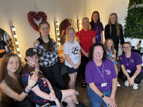 A group of adults and children smiling in a brightly lit room with vanity mirrors and floral decorations, gathered for a community event.