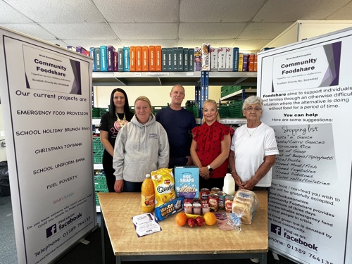 Five people standing behind a table of donated food items inside a community support centre, with banners for Community Foodshare visible in the background.