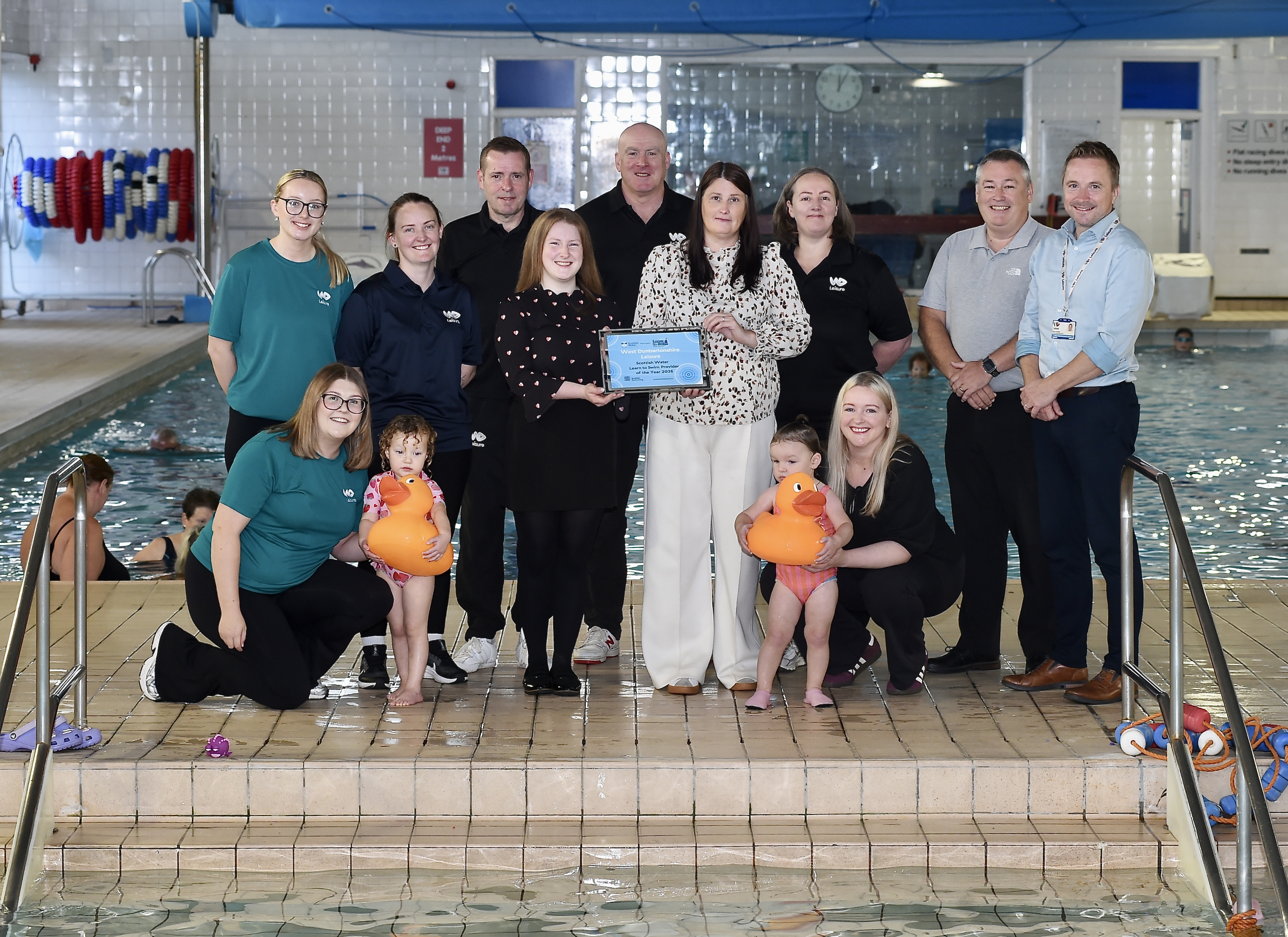 Councillors, West Dunbartonshire Leisure Trust staff and two children with yellow ducks pose with the Scottish Water Learn to Swim Provider of the Year award.